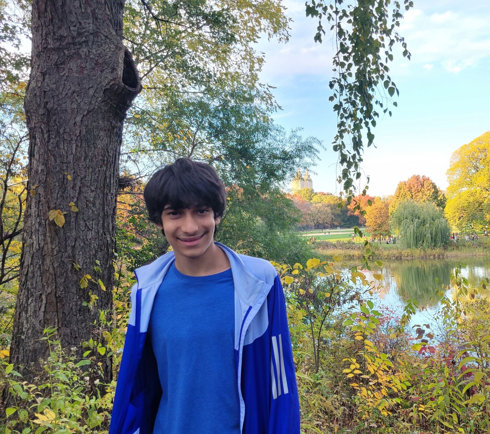 photo of a smiling Nicholas in a blue t-shirt and blue windbreaker in front of lake with trees and yellow-green flora