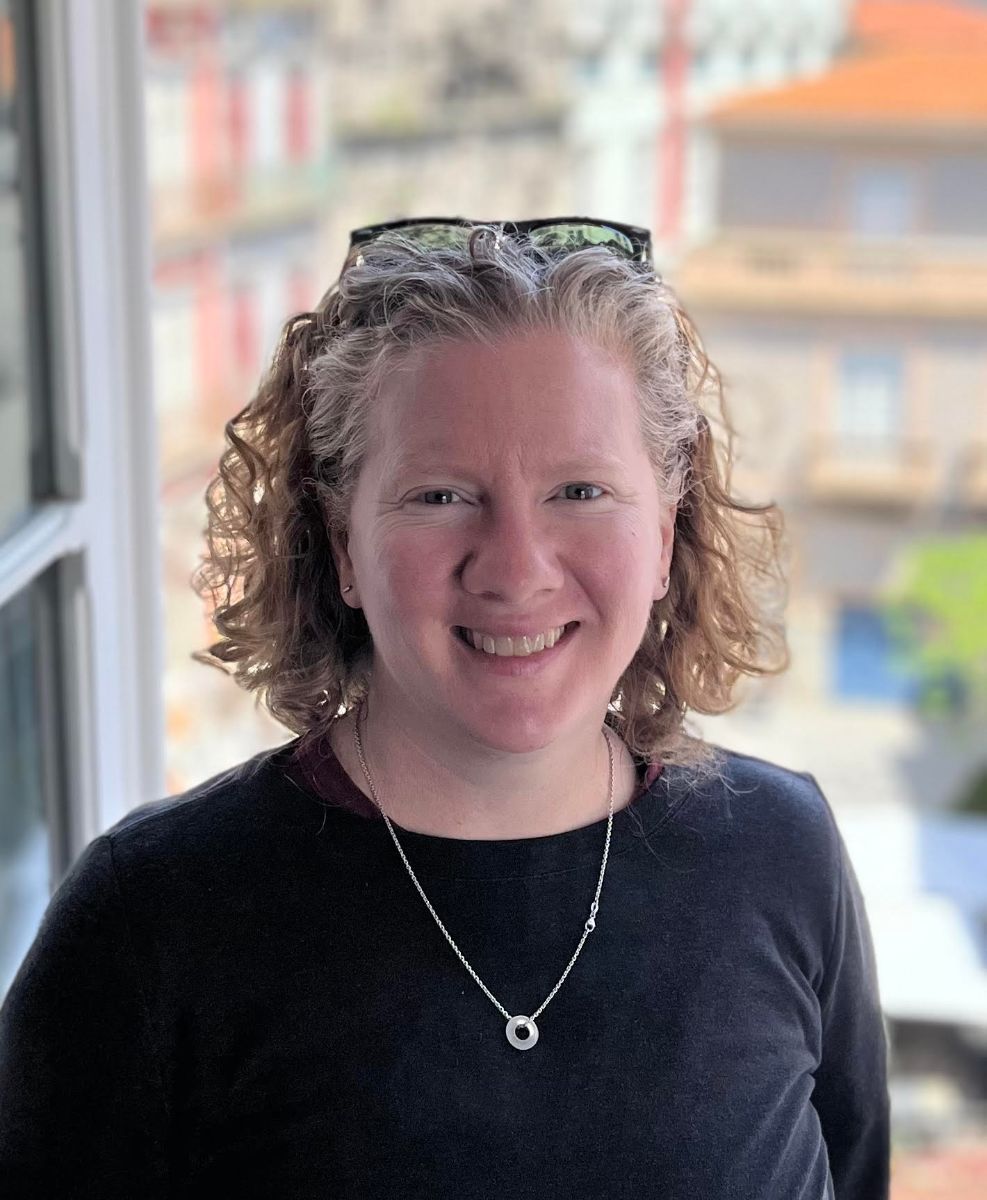 headshot of Meg Kross Lee smiling with shoulder length curly blond hair wearing a long-sleeve black top and a silver pendant necklace